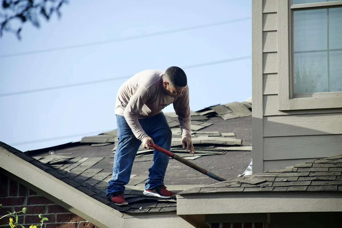 A roofer stripping old shingles from a damaged house roof